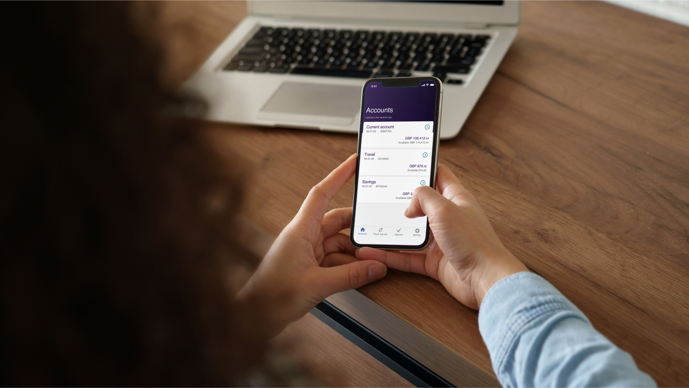 A person holding a smartphone, utilizing a mobile banking application. The “Accounts” screen is active and displays three bank accounts and their balance in GBP.