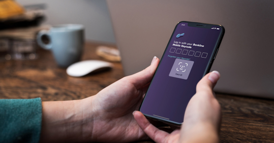 A person holding an iPhone displaying a log in screen. The FaceID functionality of iOS is displayed, together with a heading that reads “Log in with your Bankline Mobile Keycode”, followed by six empty number fields. Below there’s a text link saying “Forgotten your Keycode?”.
