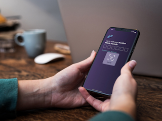 A person holding an iPhone displaying a log in screen. The FaceID functionality of iOS is displayed, together with a heading that reads “Log in with your Bankline Mobile Keycode”, followed by six empty number fields. Below there’s a text link saying “Forgotten your Keycode?”.