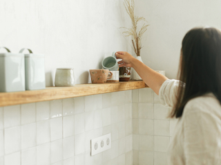 A person is seen from behind while placing a light blue ceramic cup on a wooden shelf in a minimalistic kitchen. The shelf is decorated with various ceramic mugs, jars, and a vase with dried plants. The walls have a light tone, and the tiled backsplash is white. The overall decor exudes a clean, modern, and cozy aesthetic.