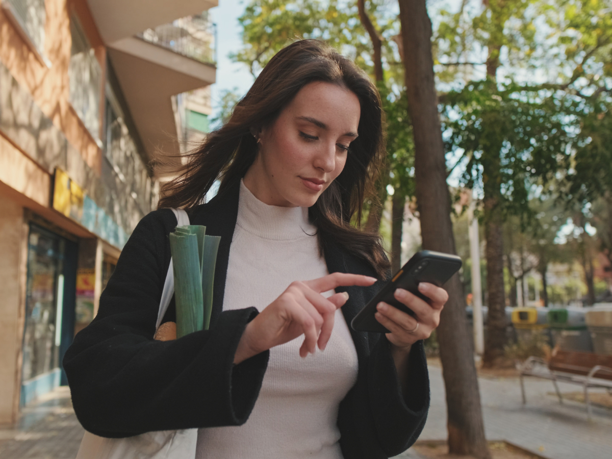 A woman carrying groceries walks outdoors while using her smartphone, surrounded by trees and buildings.