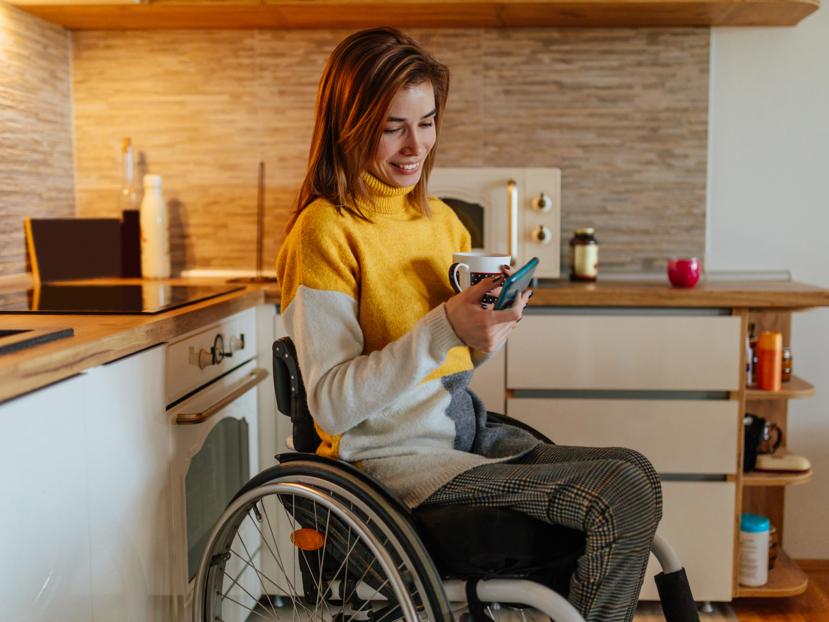 A woman sitting in a wheelchair in a modern kitchen, wearing a yellow and gray sweater, holds a coffee mug in one hand and looks at her mobile phone with a smile. The background shows kitchen countertops, cabinets, and appliances, giving a cozy and relaxed atmosphere.