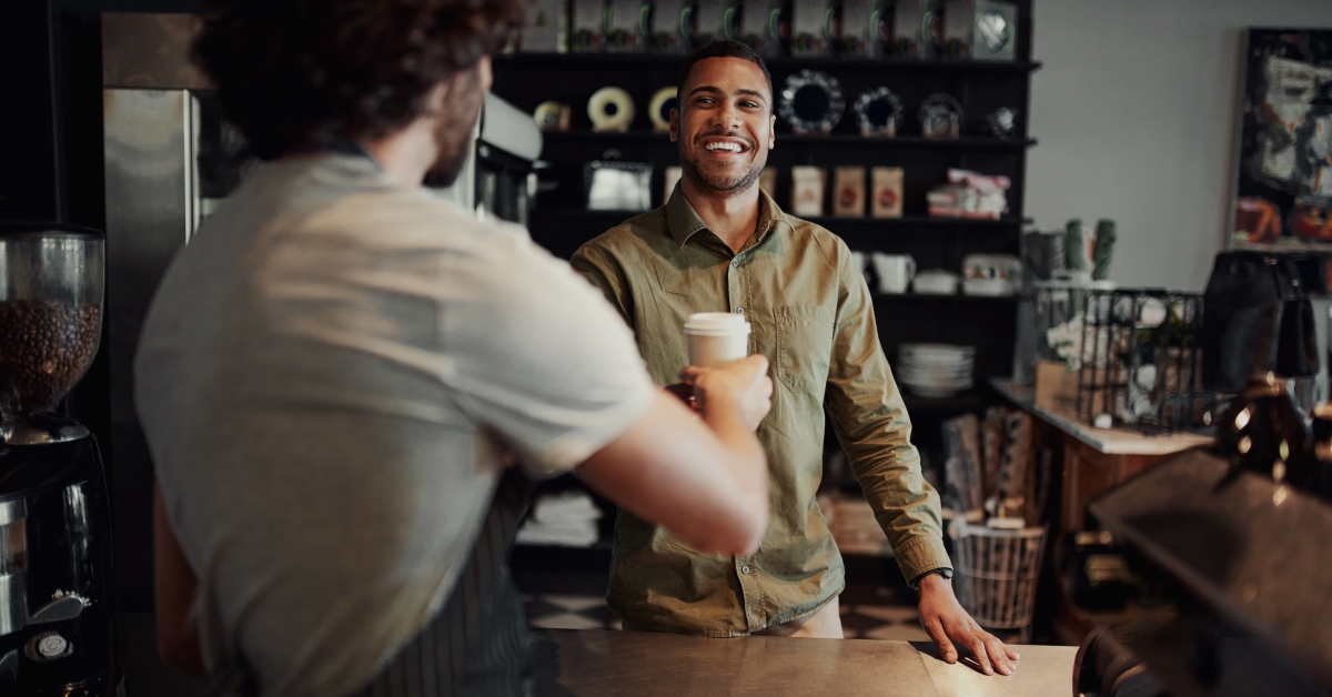 A customer at a café counter smiles as they receive a takeaway coffee cup from a barista. The customer, dressed in a light green button-up shirt, stands on the other side of the counter. The café background features shelves with various items, such as mugs and coffee bags. The atmosphere is warm and inviting.