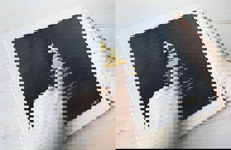 Hands holding a white tablet showing a black background with the Grammy's logo and text underneath that seems to read "The 58th Grammys", with a date and details below that are covered by the person's hand