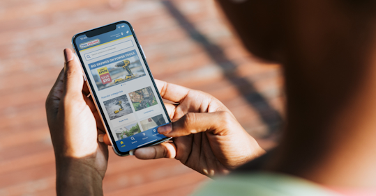 A person holding a smartphone and browsing the Toolstation website, viewing power tools and popular categories like landscaping and gardening. The screen displays a promotion for power tool savings, with a clear, user-friendly interface.