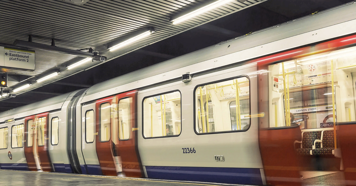 A London Underground train at Cannon Street station, captured in motion with blurred red and white doors. The train is illuminated under the station's bright lights, with empty seats visible through the windows."