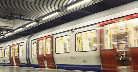 A London Underground train at Cannon Street station, captured in motion with blurred red and white doors. The train is illuminated under the station's bright lights, with empty seats visible through the windows."