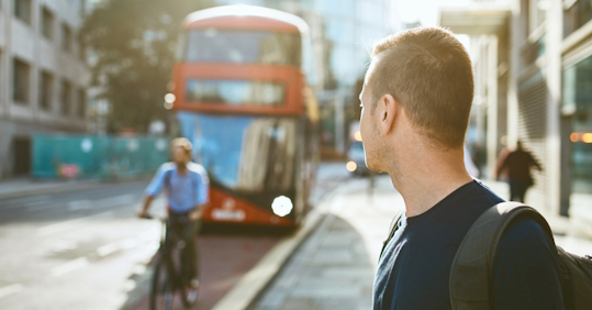 A man with a backpack stands on a city street, looking toward an approaching red double-decker bus. A cyclist rides past on the street, with blurred city buildings in the background, suggesting an urban commuting scene.