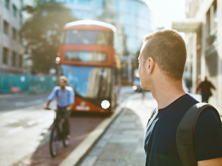 A man with a backpack stands on a city street, looking toward an approaching red double-decker bus. A cyclist rides past on the street, with blurred city buildings in the background, suggesting an urban commuting scene.