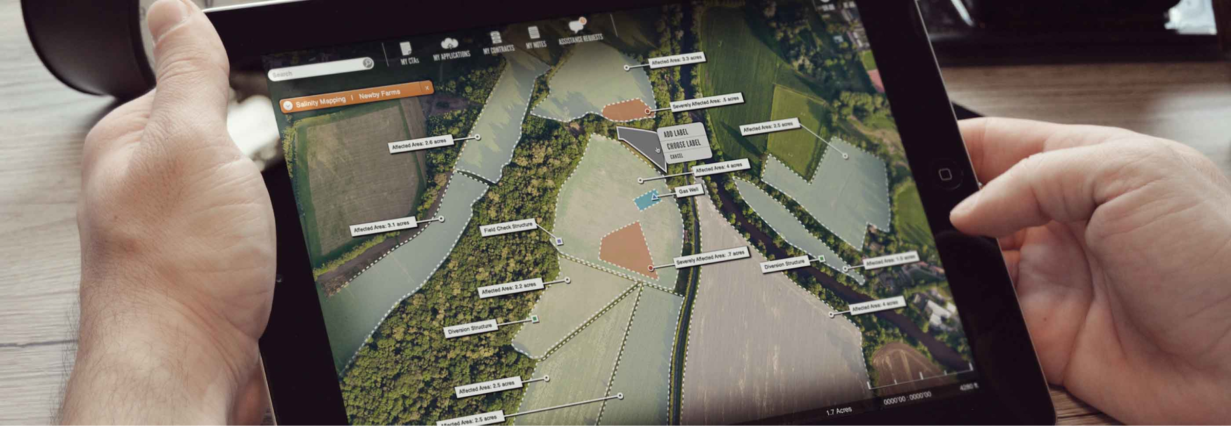 Hands holding a tablet with the interface of the web app for the U.S. Department of Agriculture, showing an aerial picture, where land is divided into different sections.