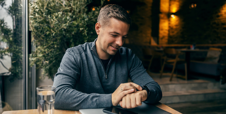 The image shows a man in casual attire looking down at his smartwatch. The background is softly blurred, suggesting an indoor or urban setting.