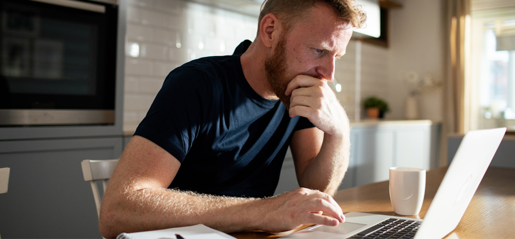 A man is sitting at a kitchen table, deeply focused on his laptop. He has a thoughtful expression, with one hand resting on his chin and the other on the keyboard. The setting is a modern kitchen with soft lighting, creating a calm atmosphere. A white coffee mug sits on the table beside him, and a notebook with a pen is visible in the corner. The image captures a moment of concentration, likely working from home or doing research online.