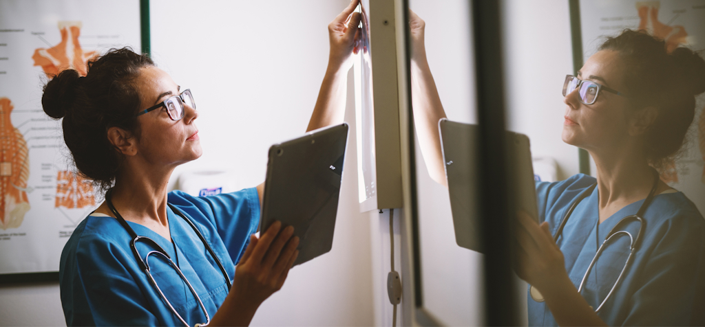 A female healthcare professional, wearing blue scrubs and a stethoscope, is carefully examining an X-ray on a lightbox while holding a tablet in her other hand. She is focused on the X-ray, studying it closely, with her reflection visible on the glass surface next to her. Behind her is a medical chart displaying anatomical diagrams. The image captures the use of both traditional and digital tools in medical diagnostics, emphasizing precision and attention to detail in healthcare settings.