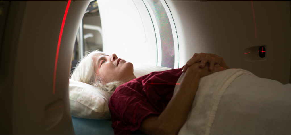 An elderly woman is lying on a bed, preparing to undergo a scan inside an MRI or CT machine. She appears calm and relaxed, with her hands resting on her abdomen and her head cushioned by a pillow. The scanner's red laser lines are visible, marking the alignment for the procedure. The setting is quiet and clinical, with the focus on the patient receiving advanced diagnostic imaging. The image emphasizes the use of modern medical technology for precise and thorough examinations.