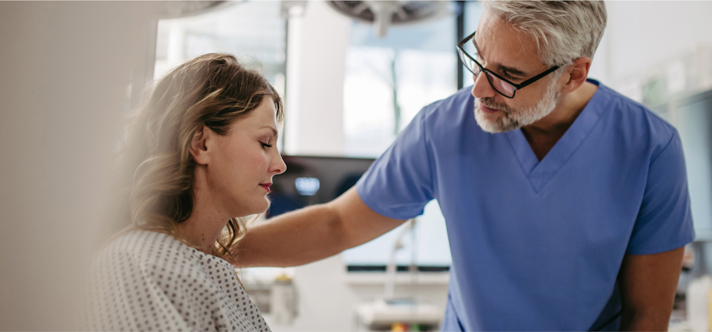 A male doctor in blue scrubs is gently comforting a female patient in a hospital gown. The doctor places a reassuring hand on the patient’s shoulder while they engage in a quiet moment. The woman, with her eyes closed, appears emotional, as if she is receiving important or difficult news. The setting is a clinical environment, with medical equipment visible in the background. The image highlights empathy and care in the doctor-patient relationship, capturing a moment of support during a vulnerable time.