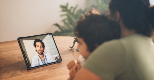 A mother and child are sitting together at a table, engaged in a video call with a doctor on a tablet. The doctor, who appears to be wearing a white coat and has a stethoscope around his neck, is speaking to them from his office, with a whiteboard visible in the background. The setting is calm and homey, with a small toy dinosaur on the table near the child. The image highlights the convenience and comfort of telemedicine, allowing patients to consult with healthcare professionals from their own home