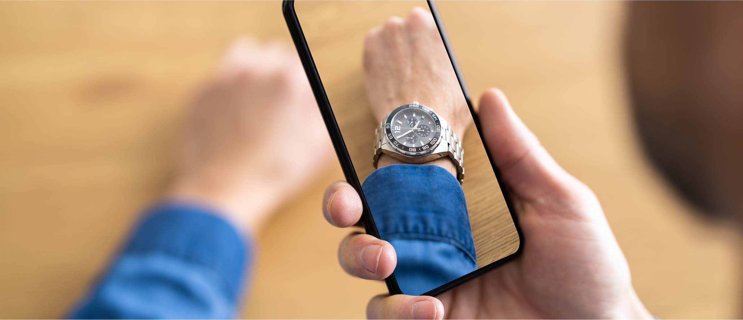 Person’s hand holding a smartphone over their wrist against a wooden table, with the phone screen showing an augmented-reality overlay of a silver watch on their wrist.