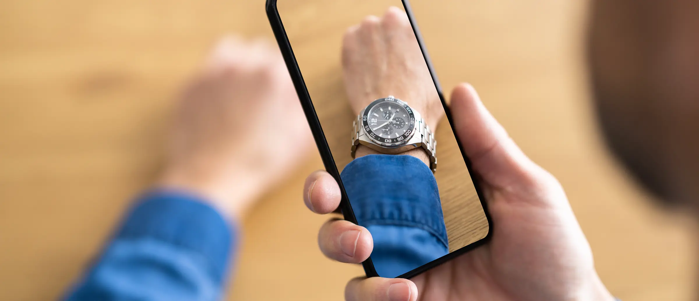 Person’s hand holding a smartphone over their wrist against a wooden table, with the phone screen showing an augmented-reality overlay of a silver watch on their wrist.
