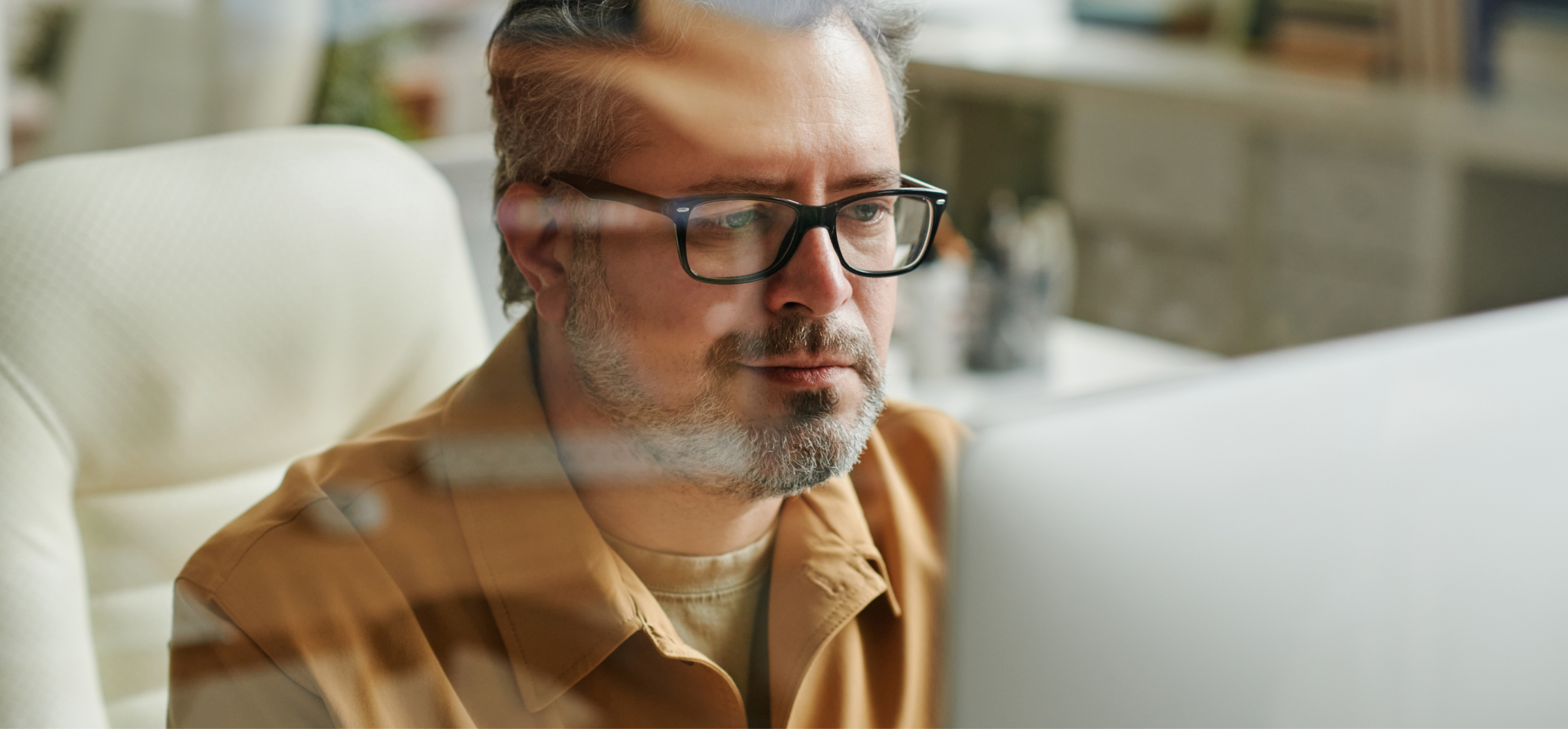 A man with glasses and a neatly trimmed beard is sitting in an office, focused on his work on a computer. He is wearing a light brown shirt and is seated in a comfortable white office chair. The lighting is soft, and the background is slightly blurred, with office supplies and shelves visible in the distance. The man appears thoughtful and concentrated, possibly analyzing data or working on a detailed project. The image captures a moment of focus and productivity in a modern office environment.
