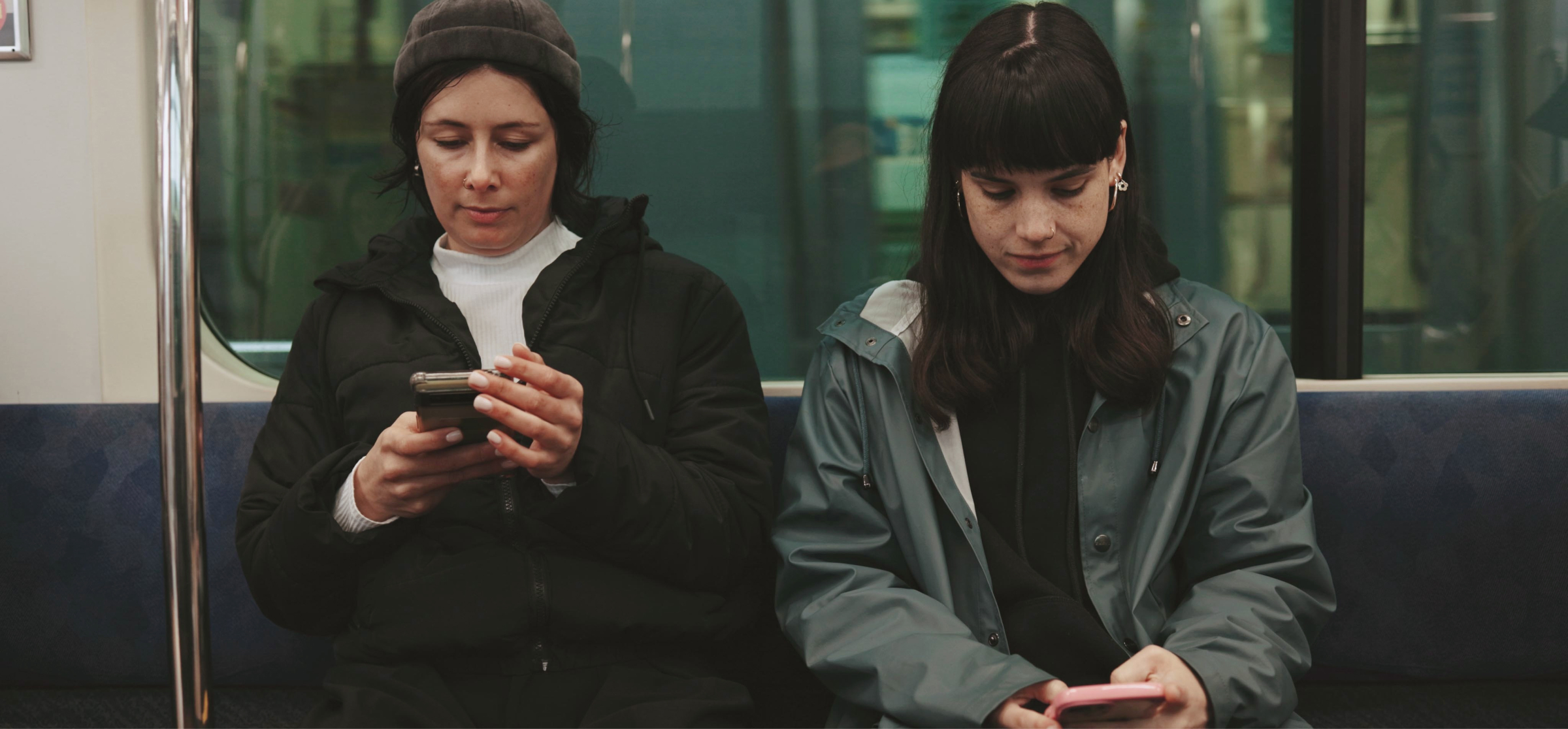  Two women are sitting side by side on public transportation, both focused on their smartphones. The woman on the left, wearing a beanie and a black jacket, is holding her phone with both hands. The woman on the right, in a teal jacket, is also engaged with her phone, looking down at the screen. The atmosphere is quiet and reflective, as both individuals are absorbed in their own devices. The setting is inside a train or subway car, with the city passing by outside the window. The image conveys the modern-day experience of commuting with technology.