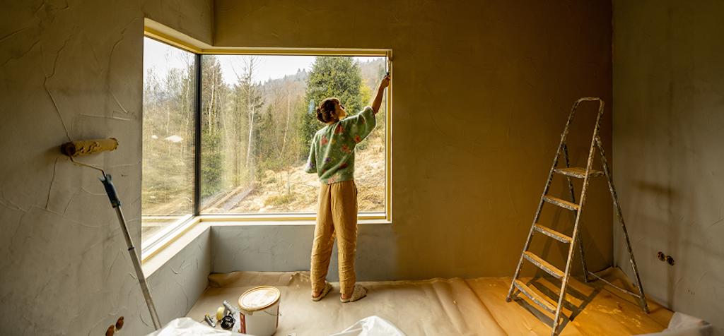 Woman painting a window frame in a sunlit room during a home improvement project, featuring DIY renovation and interior design updates.