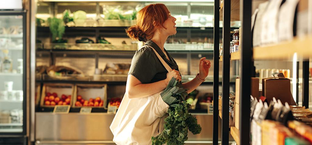 Woman holding reusable bag and fresh produce while browsing shelves in a well-stocked supermarket, highlighting sustainable grocery shopping and healthy living.