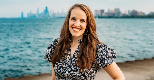 Kerry Penny, SVP and Global Head of Brand & Content at Valtech, stands near a waterfront with a blurred city skyline in the background. She is wearing a navy blue floral dress with puffed sleeves and gold hoop earrings. The weather appears clear, and the water behind her is calm.