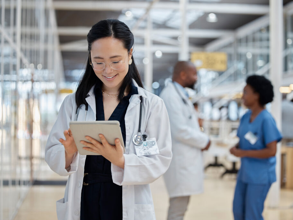 Female doctor wearing a white lab coat and glasses, smiling while using a tablet in a bright, modern healthcare facility, with medical staff working or chatting in the background