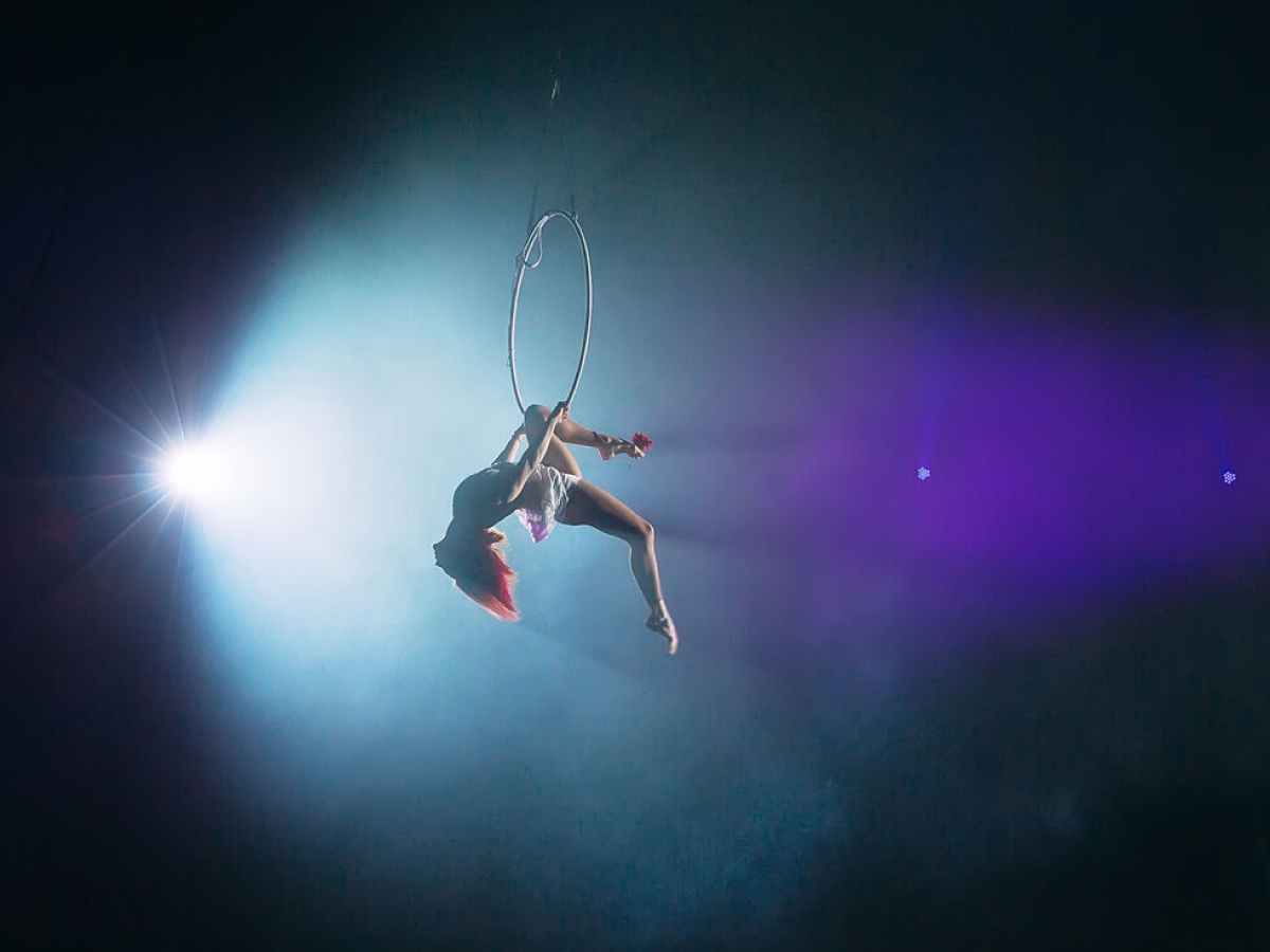 Aerial acrobat performing on a suspended hoop under dramatic stage lighting with blue and purple hues. The performer is mid-movement, displaying strength and elegance in a gravity-defying pose.