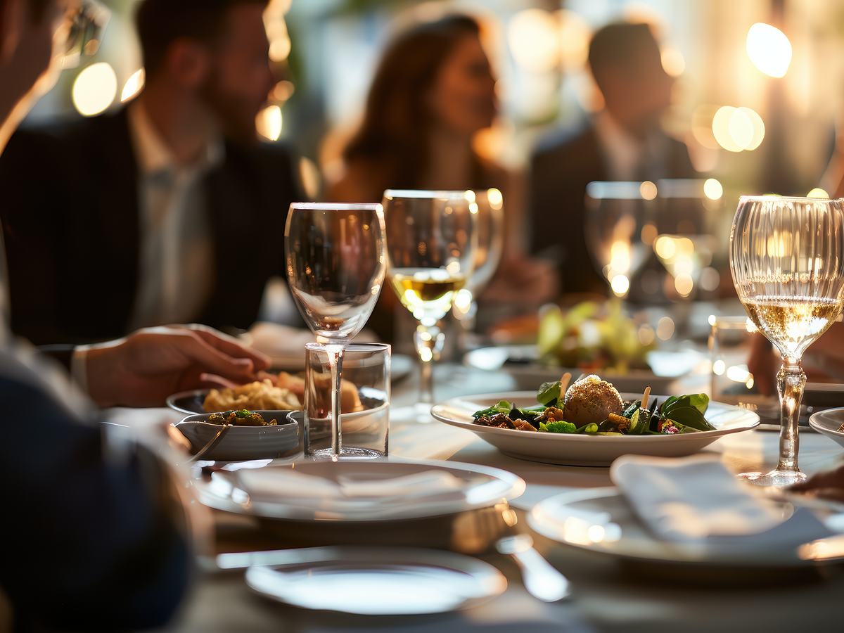 Close-up of a sophisticated dinner table during a business event, featuring wine glasses, gourmet dishes, and elegantly dressed people engaged in conversation in a warmly lit setting.