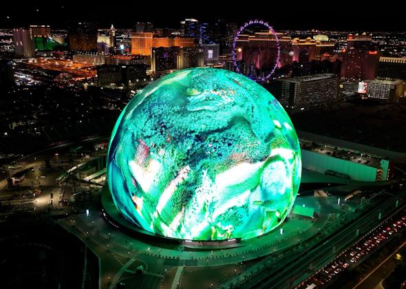A stunning nighttime aerial view of the Las Vegas Sphere, illuminated with vibrant green and white digital visuals. The city skyline, including hotels, casinos, and the High Roller Ferris wheel, is visible in the background, showcasing the bright lights of the Las Vegas Strip. The futuristic display on the Sphere highlights cutting-edge technology and immersive digital experiences.