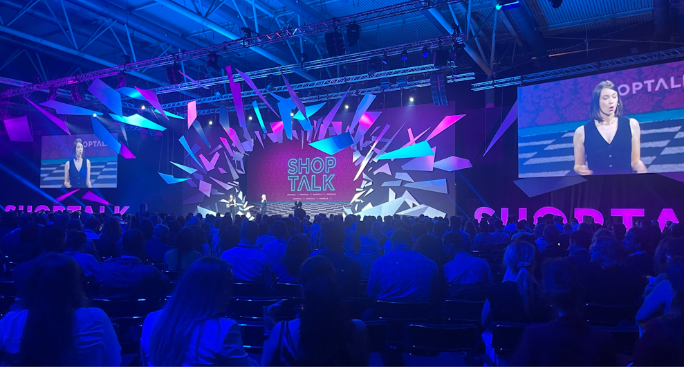 A large audience attends the Shoptalk conference keynote, featuring a dynamic stage design with bright pink and blue lighting. A speaker is presenting on stage, with her image displayed on two large screens.