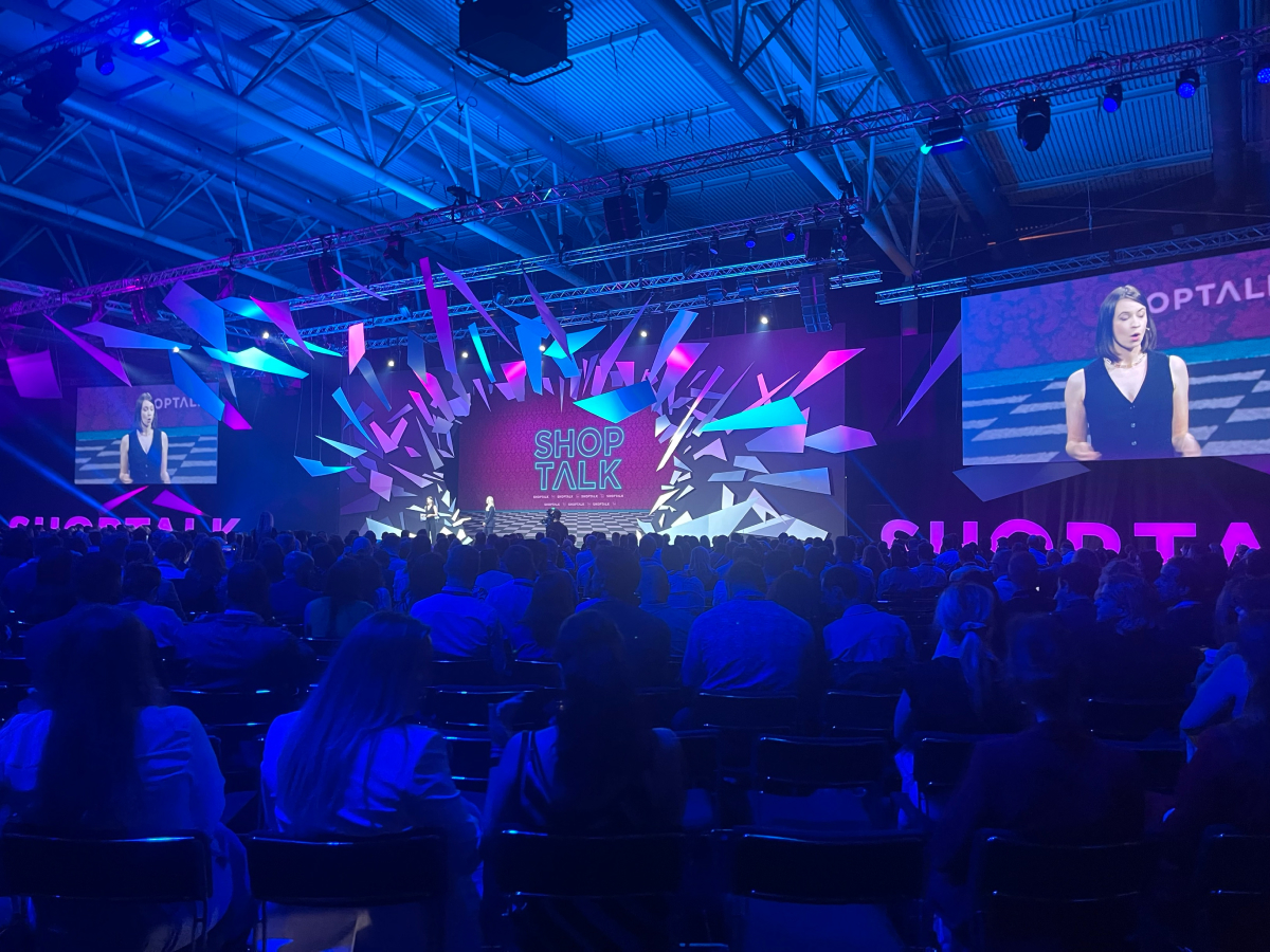 A large audience attends the Shoptalk conference keynote, featuring a dynamic stage design with bright pink and blue lighting. A speaker is presenting on stage, with her image displayed on two large screens.