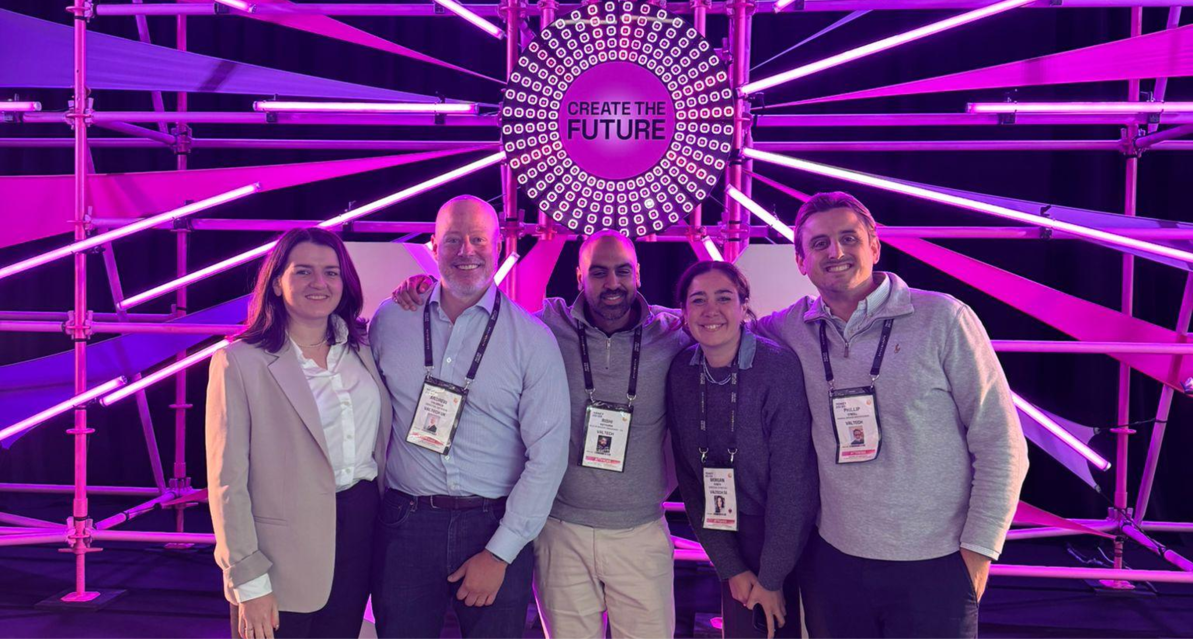 Five conference-goers stand side by side, smiling, with arms around each other in front of a vibrant, purple-lit backdrop made of intersecting light bars and a circular sign that reads “CREATE THE FUTURE.” All wear event lanyards; the two on the ends wear light-colored jackets, while the three in the middle wear collared shirts and sweaters.