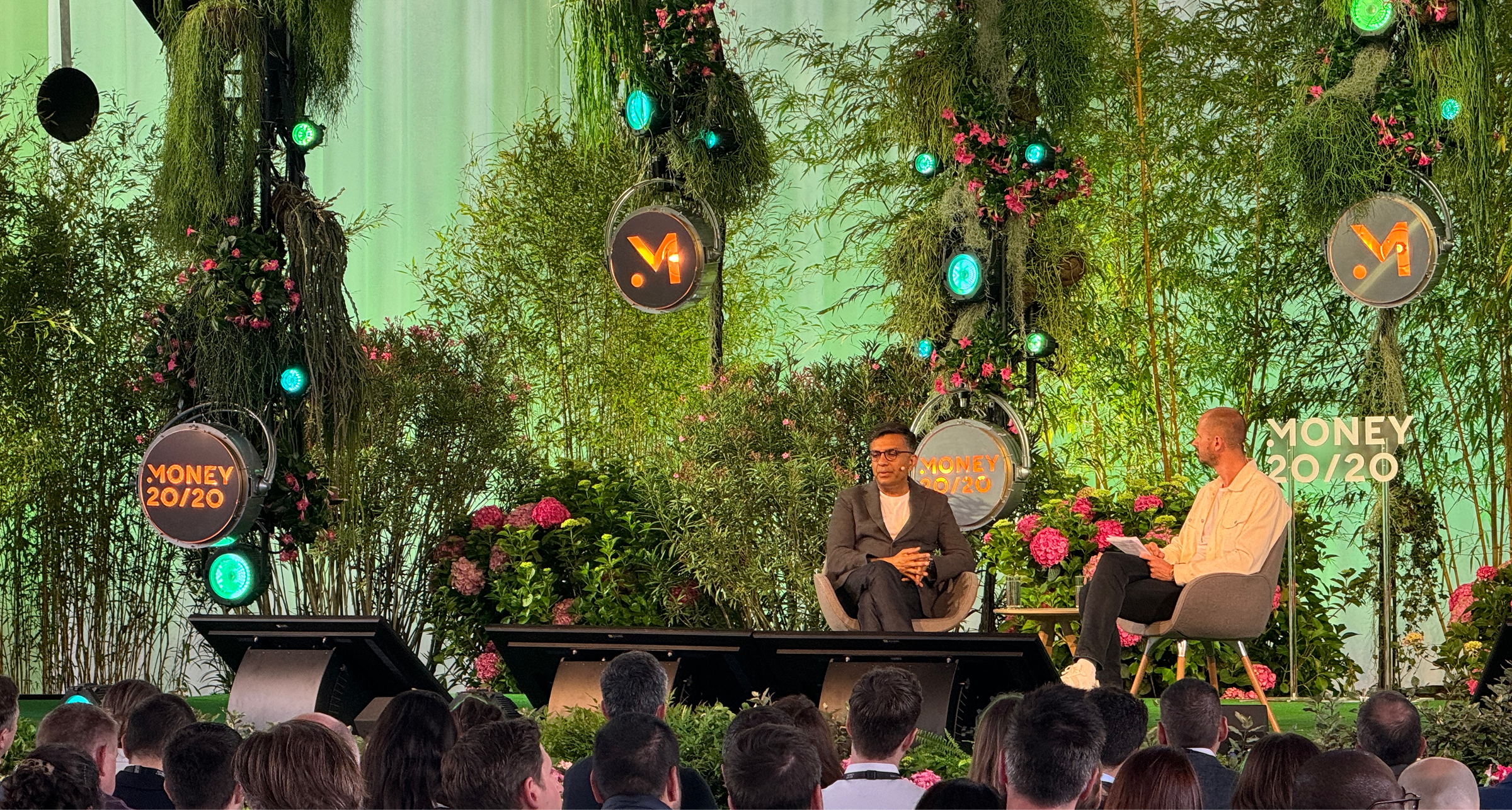 Two speakers sit facing each other on a lush, plant-filled stage dotted with pink hydrangeas and glowing circular “Money 20/20” logos. The speaker on the left wears a dark suit and glasses with hands clasped, while the speaker on the right, in a light shirt, holds notes. Audience members watch from the foreground beneath low video monitors.