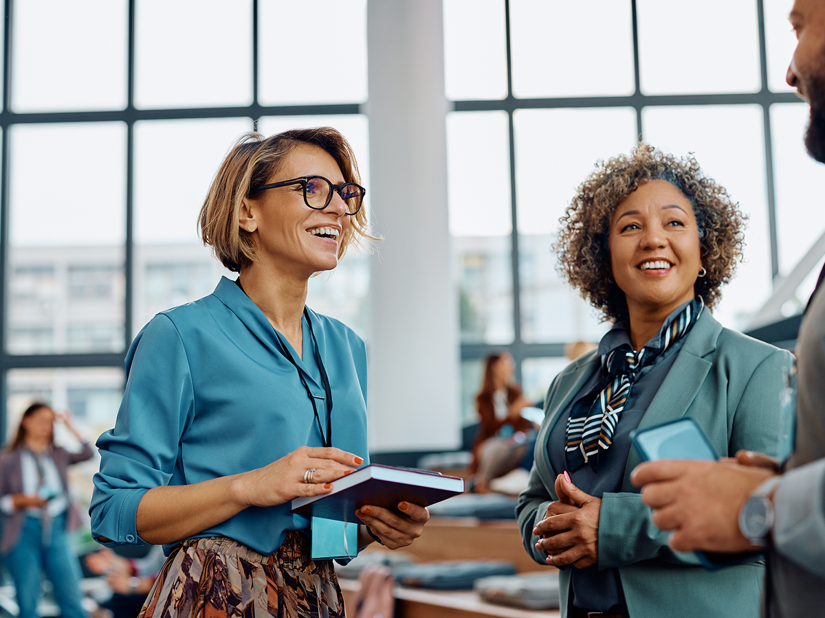 Three professionals engaging in a lively conversation at a networking event, with two women smiling and holding a notebook and phone, and a modern office background with large windows.