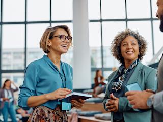 Three professionals engaging in a lively conversation at a networking event, with two women smiling and holding a notebook and phone, and a modern office background with large windows.