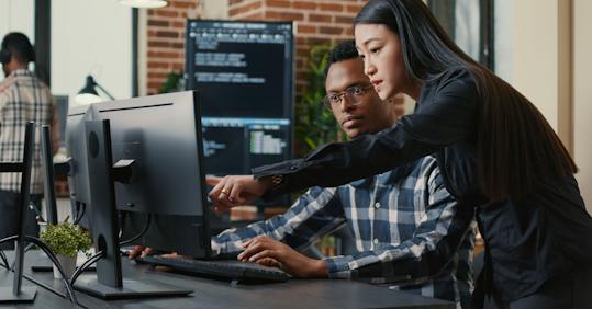 A female software developer in a black blouse is pointing at a computer screen while guiding a male colleague in a checkered shirt at a modern tech office. The screen displays lines of code, indicating a software development or programming task. The background features additional team members working in a collaborative workspace with exposed brick walls.