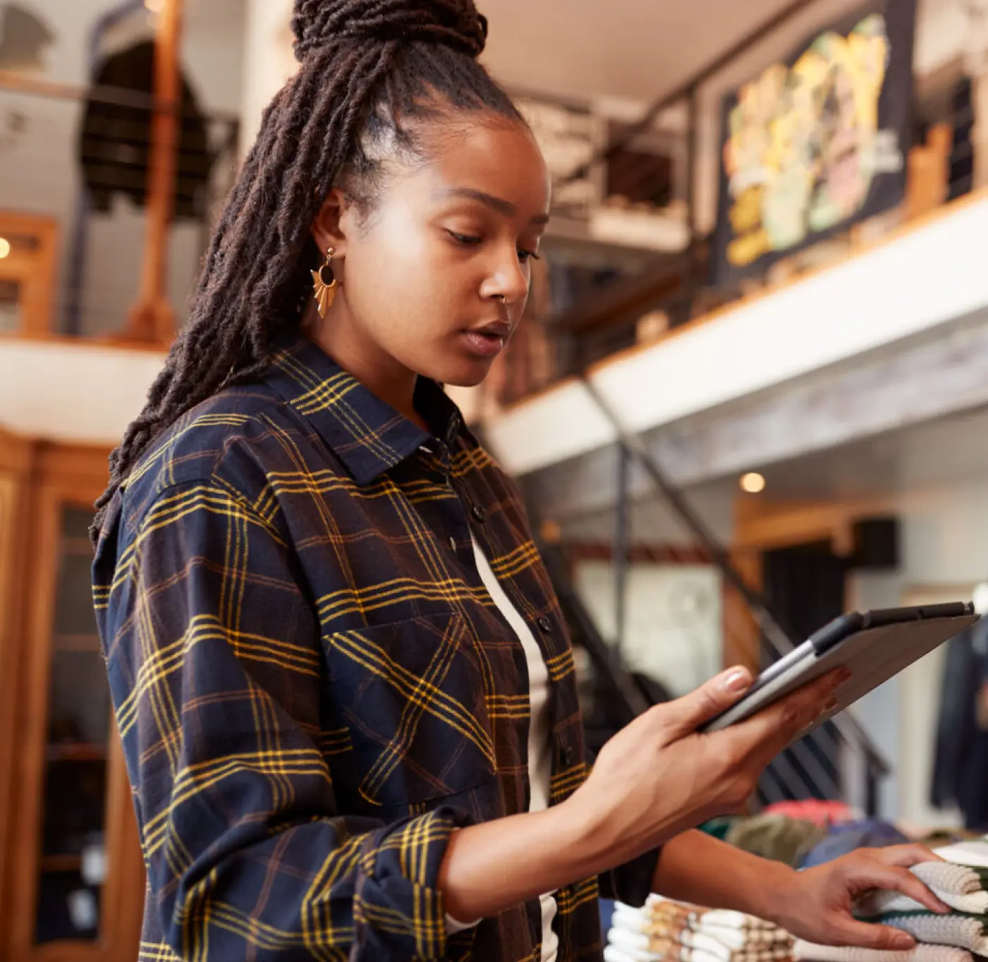 A young female retail worker in a plaid shirt is using a tablet to check inventory in a clothing store. She is standing near a neatly stacked display of folded sweaters, reviewing stock or processing sales data. The store has a stylish, rustic interior with wooden beams, clothing racks, and an upper-level loft, creating a boutique shopping atmosphere.
