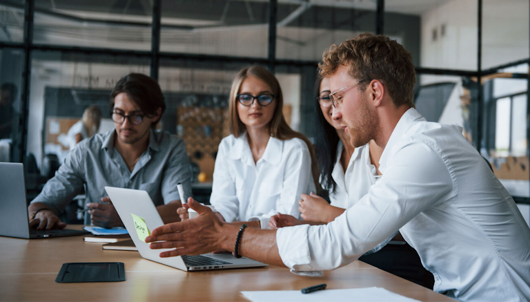 A group of four young professionals in business casual attire are engaged in a discussion around a table in a modern office. One man in a white shirt is actively presenting an idea while gesturing towards a laptop. The other team members, including two women in white blouses and a man in glasses, are attentively listening. The workspace has glass partitions and an open-concept design, indicating a collaborative and creative work environment.