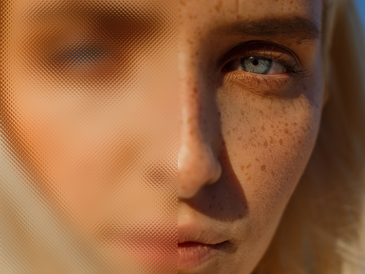 Close-up of a person's face, half obscured by textured glass, with freckles and a blue eye visible