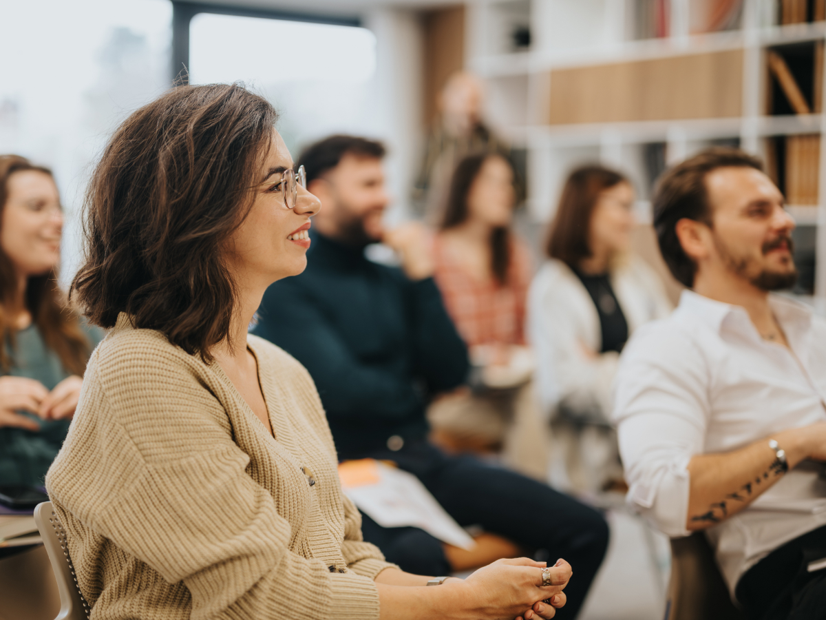 A group of people is sitting in a classroom or conference setting, engaged and smiling as they listen to a speaker. The focus is on a woman in the foreground, who has shoulder-length brown hair and is wearing glasses and a beige cardigan.
