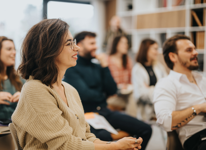 A group of people is sitting in a classroom or conference setting, engaged and smiling as they listen to a speaker. The focus is on a woman in the foreground, who has shoulder-length brown hair and is wearing glasses and a beige cardigan.