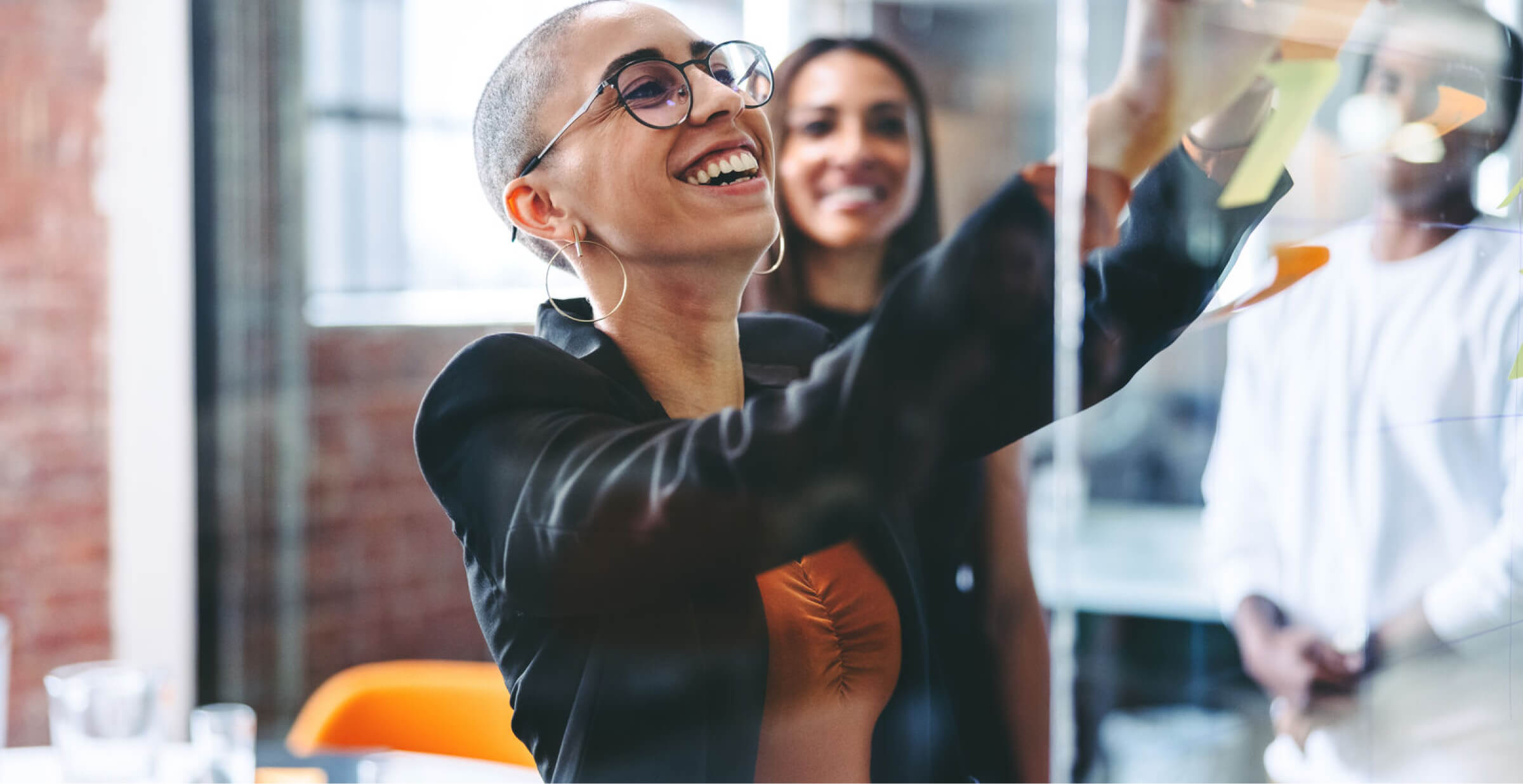 A woman with a shaved head and large hoop earrings is smiling brightly as she writes or places sticky notes on a transparent board during a collaborative session.