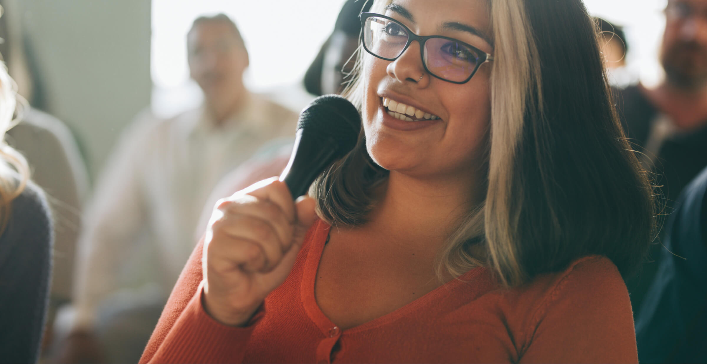 A woman with glasses and a distinctive streak of light-colored hair is holding a microphone and smiling as she speaks. 