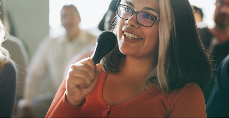 A woman with glasses and a distinctive streak of light-colored hair is holding a microphone and smiling as she speaks.