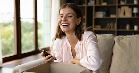 A woman smiles joyfully while holding her phone and unboxing a package, having ordered it online on a commerce website