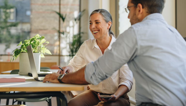 "Two colleagues collaborating in a bright, modern office setting, with one smiling while engaging with a laptop and a plant in the background.