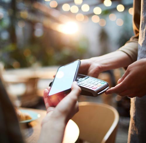 A close-up of a person making a contactless payment with a smartphone on a card reader, set against a blurred background with warm lighting and bokeh effects.