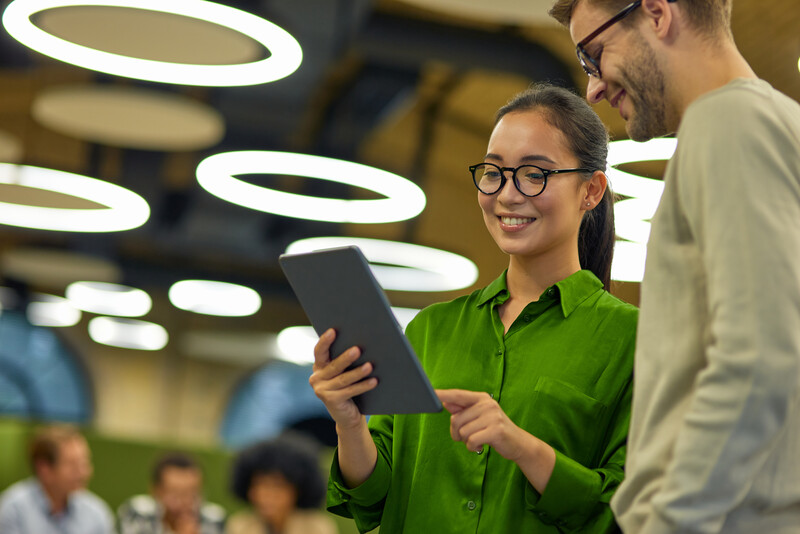 A smiling woman in a green blouse holds a tablet while standing next to a man in glasses and a light sweater, both looking at the screen together in a modern office space with circular overhead lights and a blurred group working in the background.